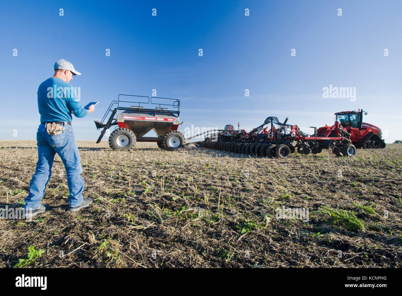 farmer using a tablet in front of a tractor and air seeder planting winter wheat in a zero till field containing canola stubble, near Lorette, Manitoba, Canada Stock Photo