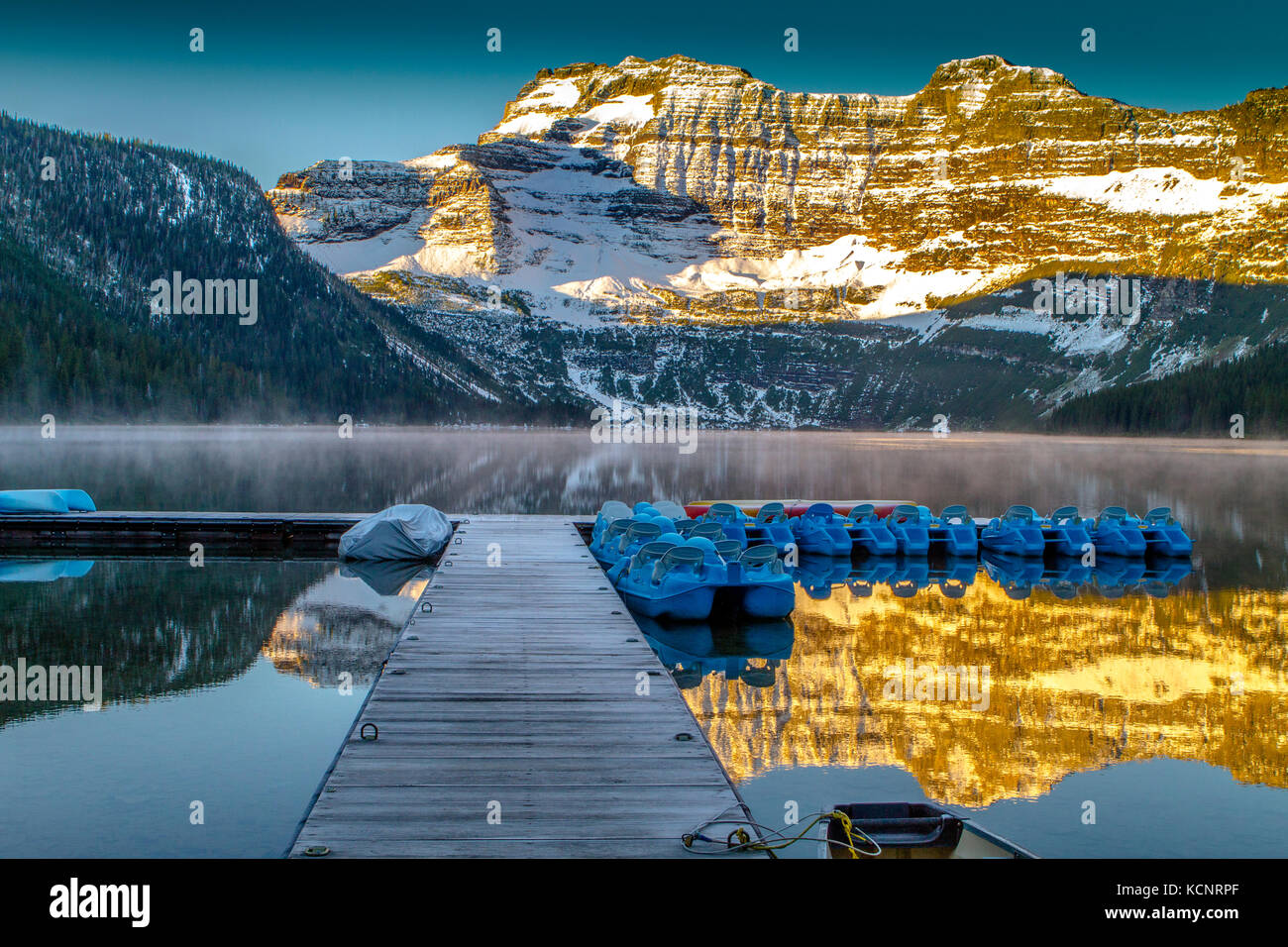 Cameron Lake in the early morning light showing wooden pier and canoe, with mountain reflections in lake. Waterton National Park, Alberta, Canada Stock Photo