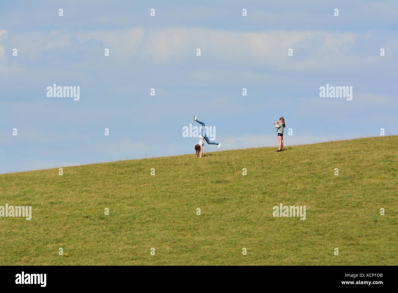 Young girl does a cartwheel on a hill top whilst her friend takes her picture on her smartphone at Barton hills nature reserve, Bedfordshire, UK Stock Photo