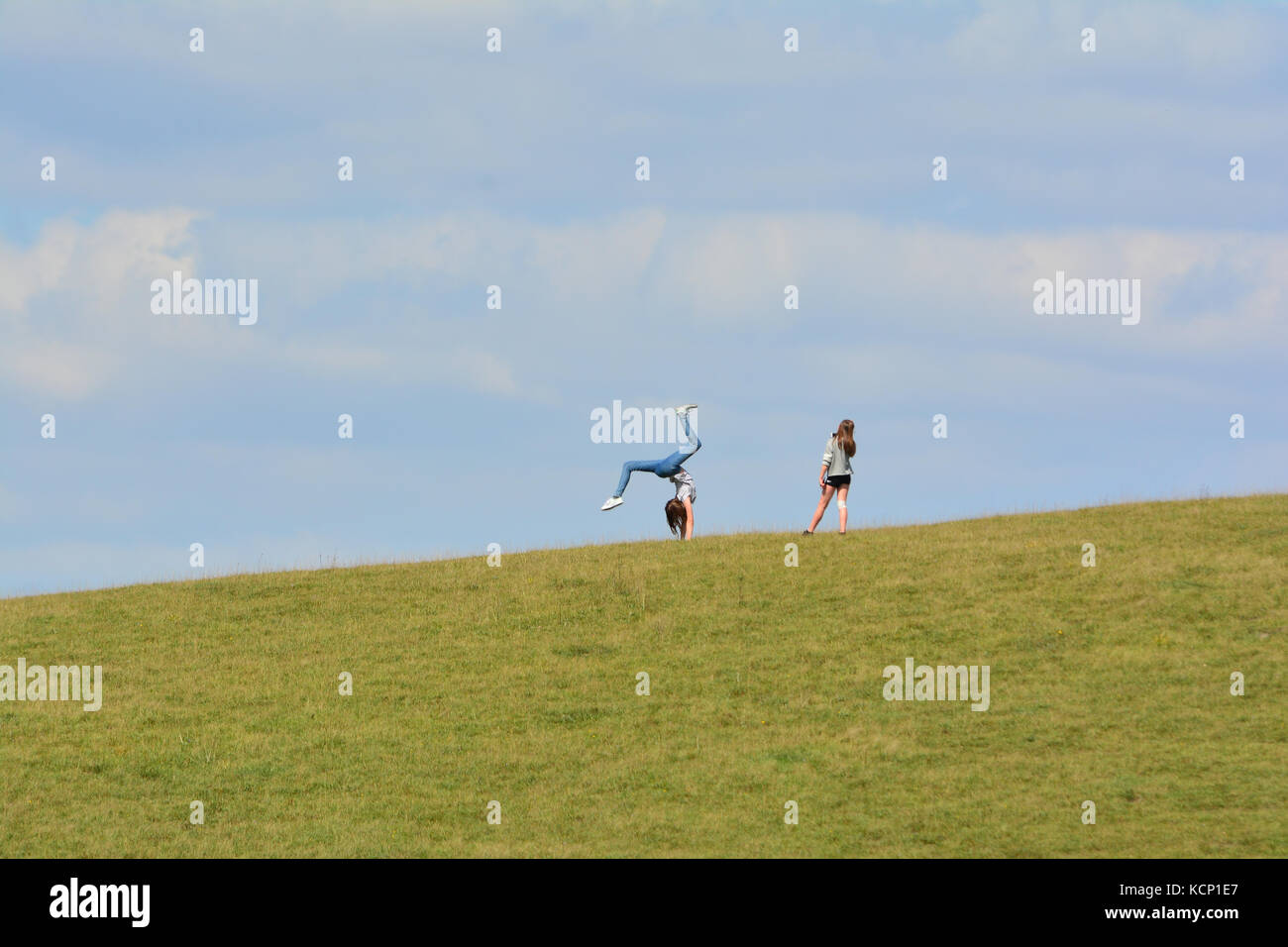 Young girl does a cartwheel on a hill top whilst her friend takes her picture on her smartphone at Barton hills nature reserve, Bedfordshire, UK Stock Photo