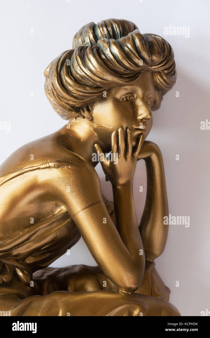 part of gold coloured ornament of woman with chin in hands and elbows resting on knees set on white background Stock Photo