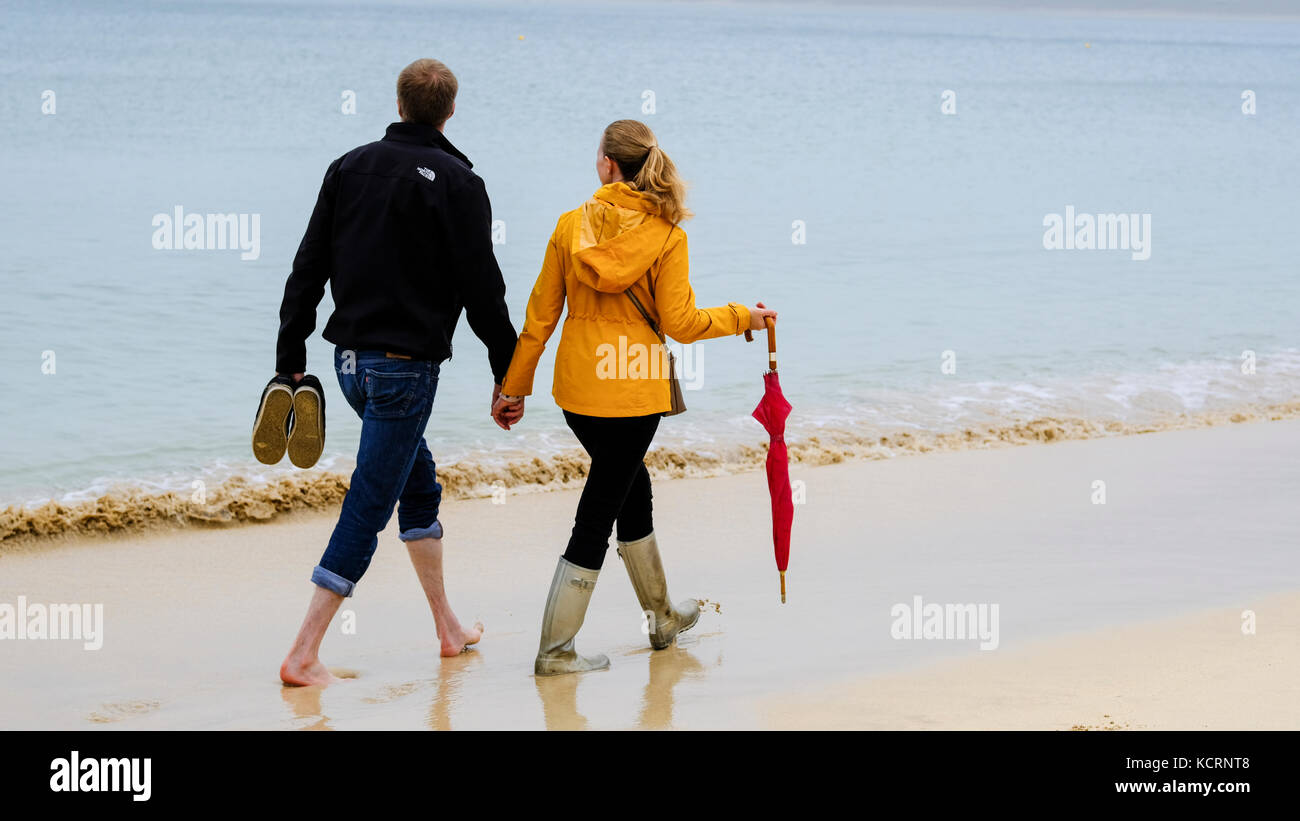 Coupler walking on Porthminster beach holding hands. Man walking barefoot while woman weras wellington boots and carries umbrella. Stock Photo