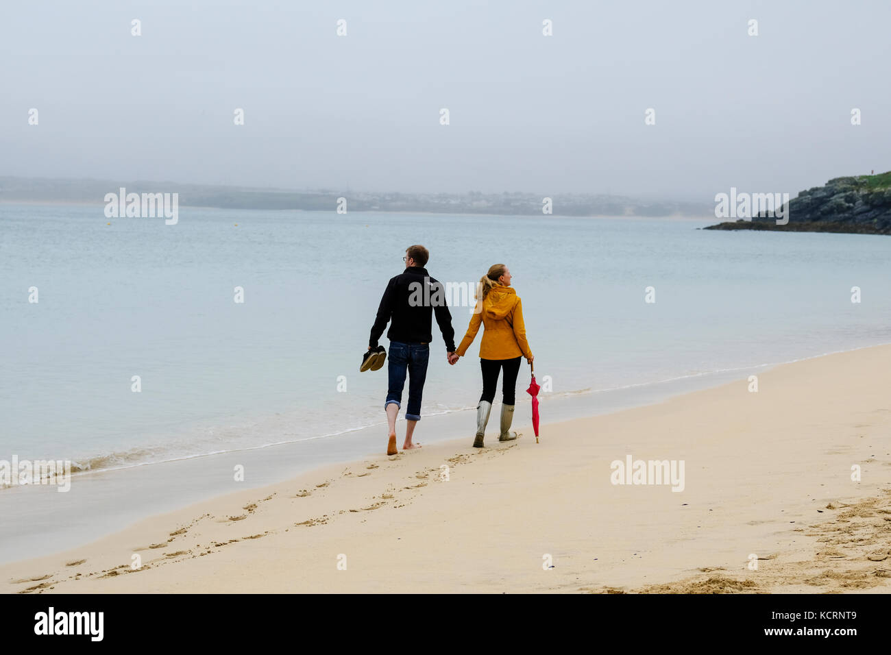 Coupler walking on Porthminster beach holding hands. Man walking barefoot while woman weras wellington boots and carries umbrella. Stock Photo