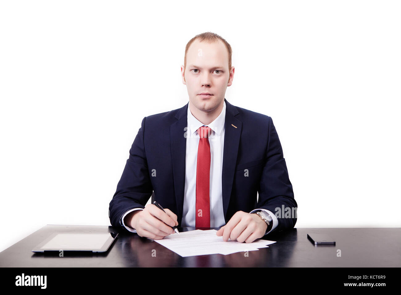 Portrait of confident young realtor in suit sitting at desk in office, signing contract, looking straight at desk, tablet, phone, on white background. Stock Photo