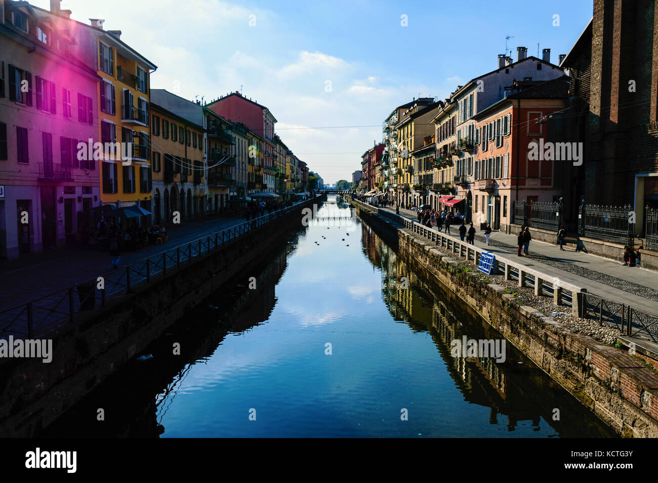 Scenic View Of The Crowded Naviglio Grande District In Milan With Water Reflections And Purple Sun Flare Against Blue Sky Stock Photo