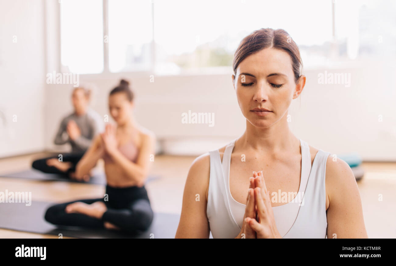 Healthy woman doing yoga in gym class with people in background. People meditating at health club. Stock Photo
