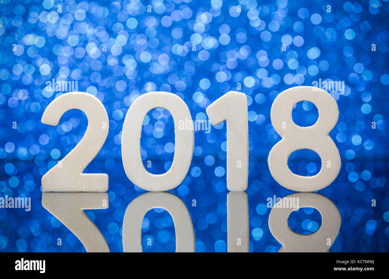 New year 2018 wood numbers reflexion on glass table in front of white lights bokeh over blue background. Stock Photo