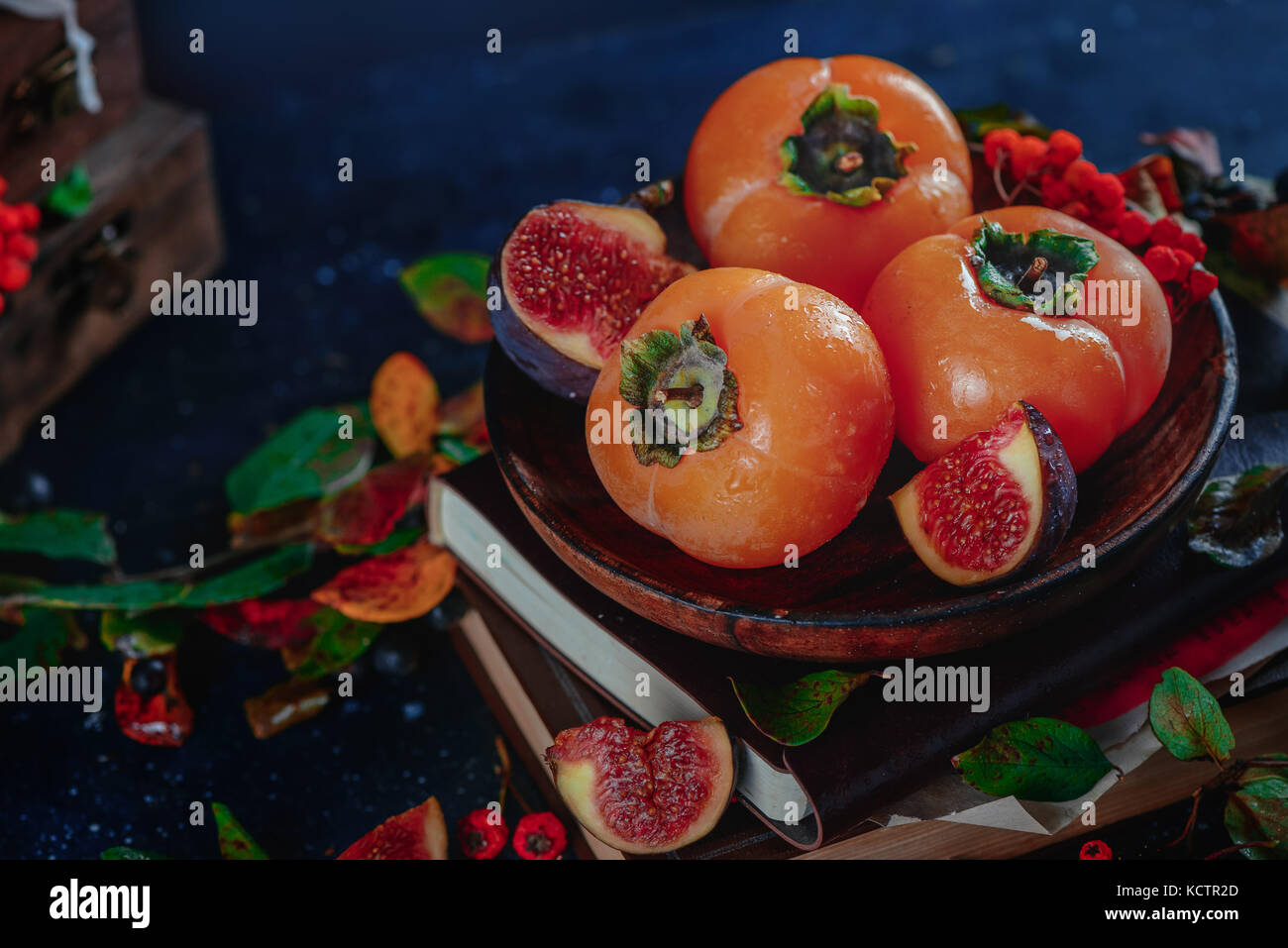 Ripe persimmons on a stack of books in a dark autumn still life with fruits and berries. Harvest concept. Dark food photography with copy space. Stock Photo