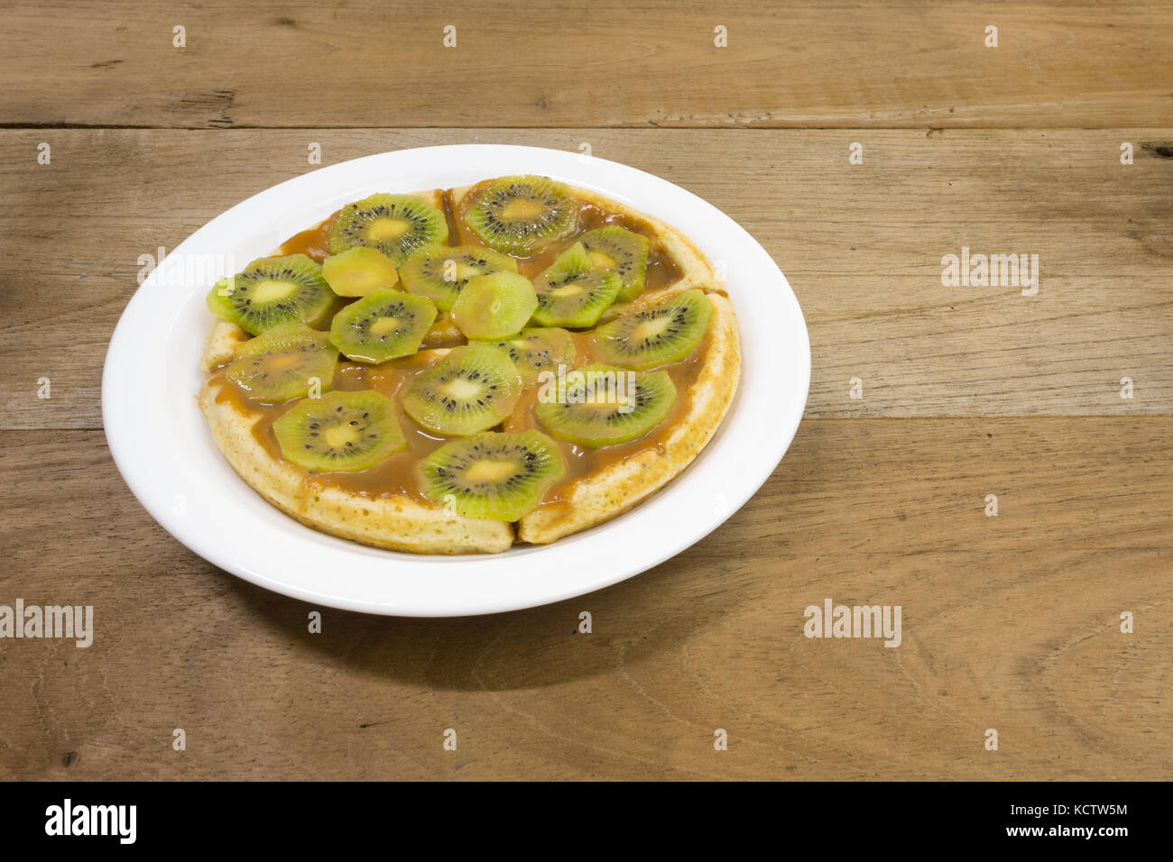 Waffle with milk caramel spread (Spanish: Dulce de leche) and kiwi fruit slices on white plate, on wooden background Stock Photo