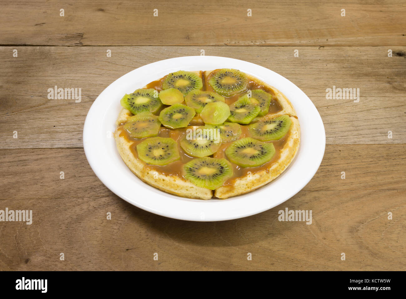 Waffle with milk caramel spread (Spanish: Dulce de leche) and kiwi fruit slices on white plate, on wooden background Stock Photo