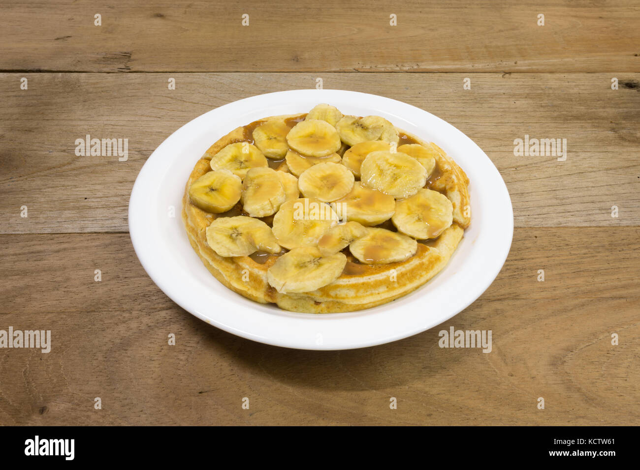 Waffle with milk caramel spread (Spanish: Dulce de leche) and banana slices on white plate, on wooden background Stock Photo
