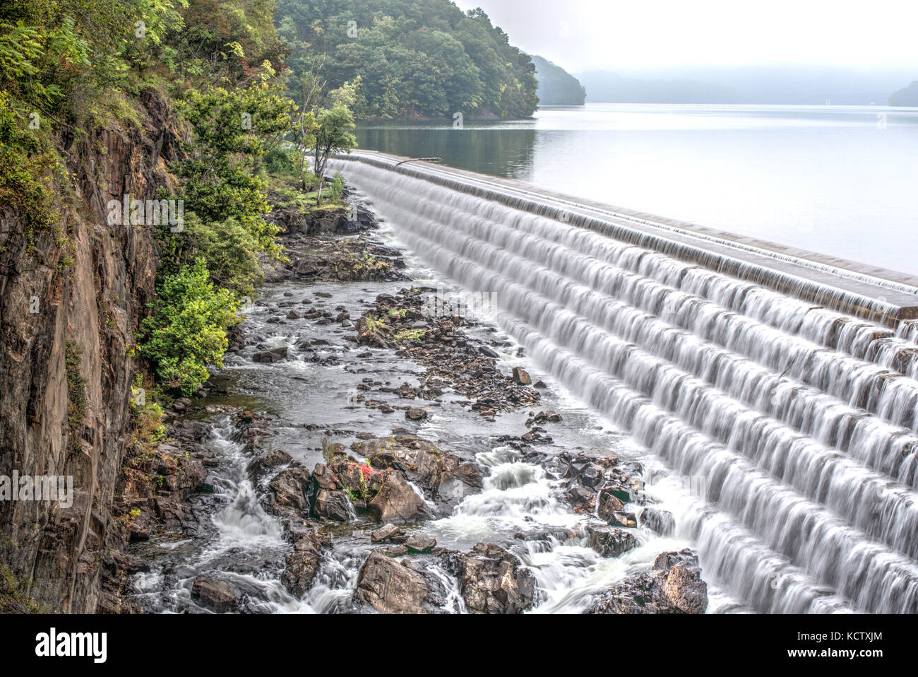 Man made spillway on a lake next to a natural cliff creates a beautiful waterfall from stepped rocks and bricks Stock Photo