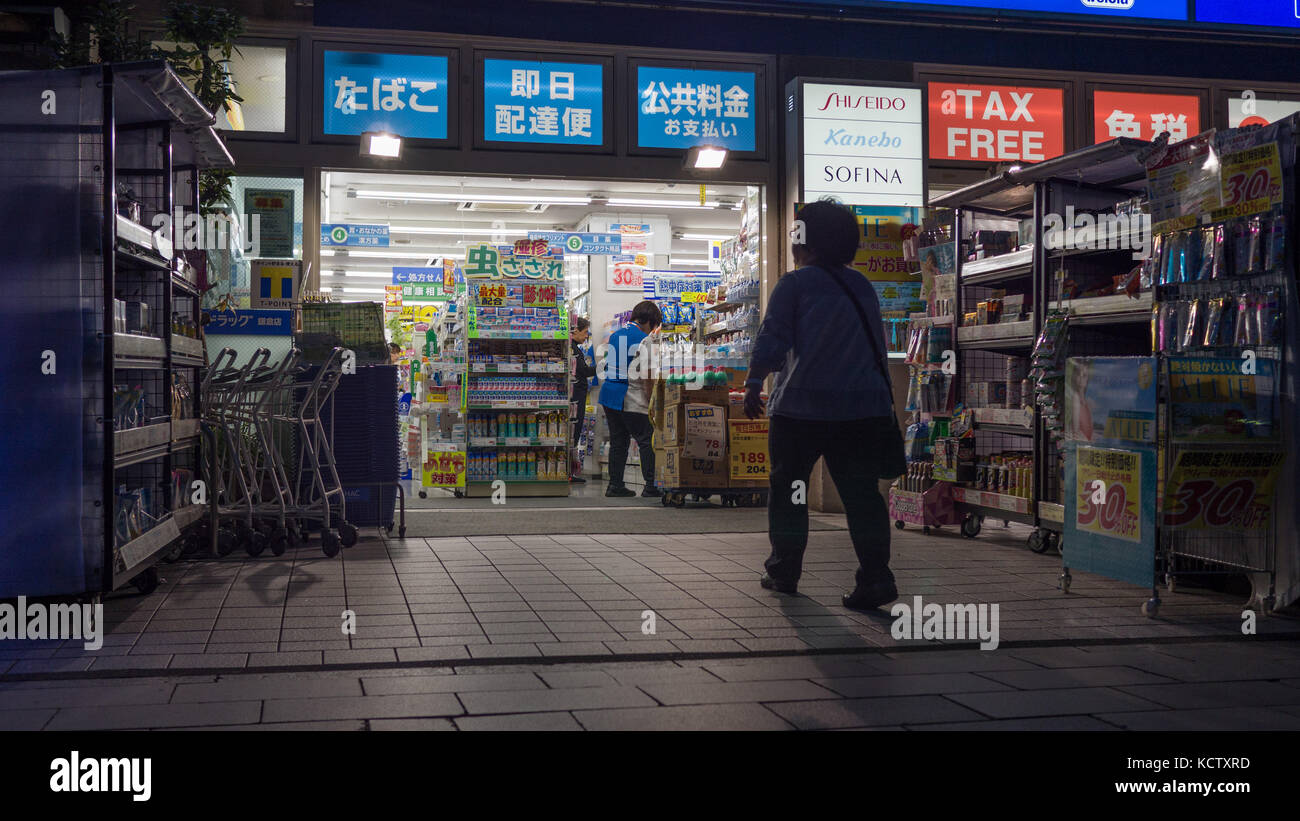 Elderly japanese woman entering a pharmacy at night. Stor lights ...