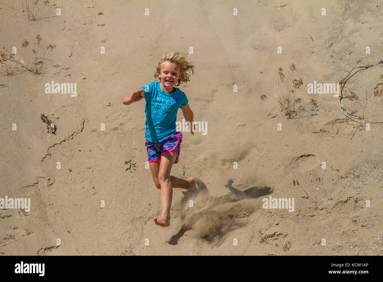 Young blonde girl, running down sand hill, laughing, hair and sand flying, having fun. Cranbrook, BC, Canada Stock Photo