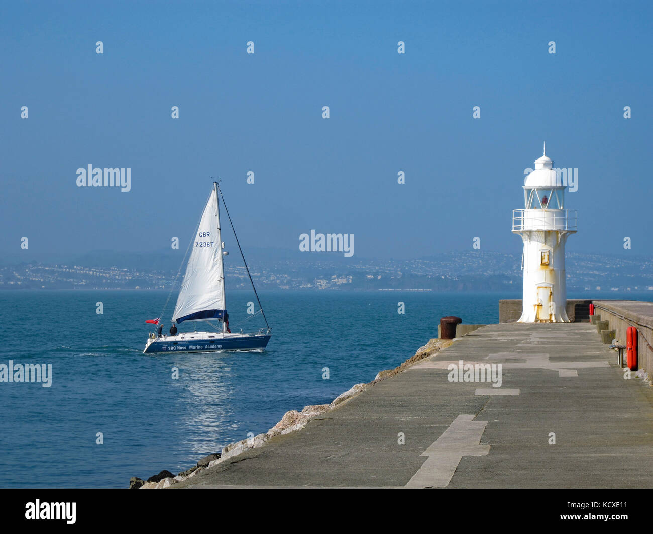 Sailboat, yacht passing the lighthouse on the end of the breakwater at Brixham, Torbay, English Riviera, Devon, UK. Summer. Copy space. Seafood Coast Stock Photo