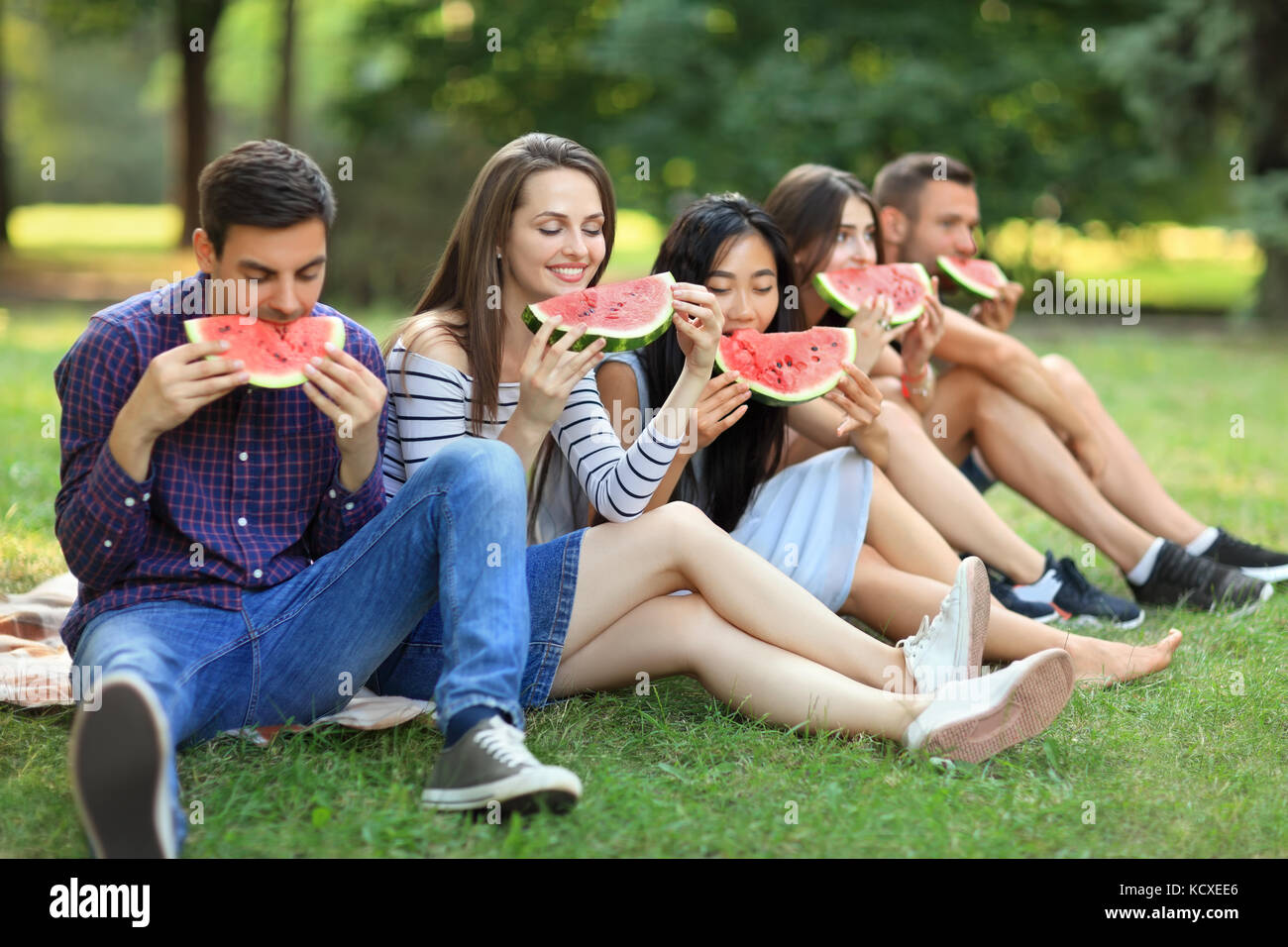 Five beautiful young people eating juicy ripe watermelon outdoors. Happy friends sitting on grass and bite off piece of fruit with pleasure. Healthy l Stock Photo