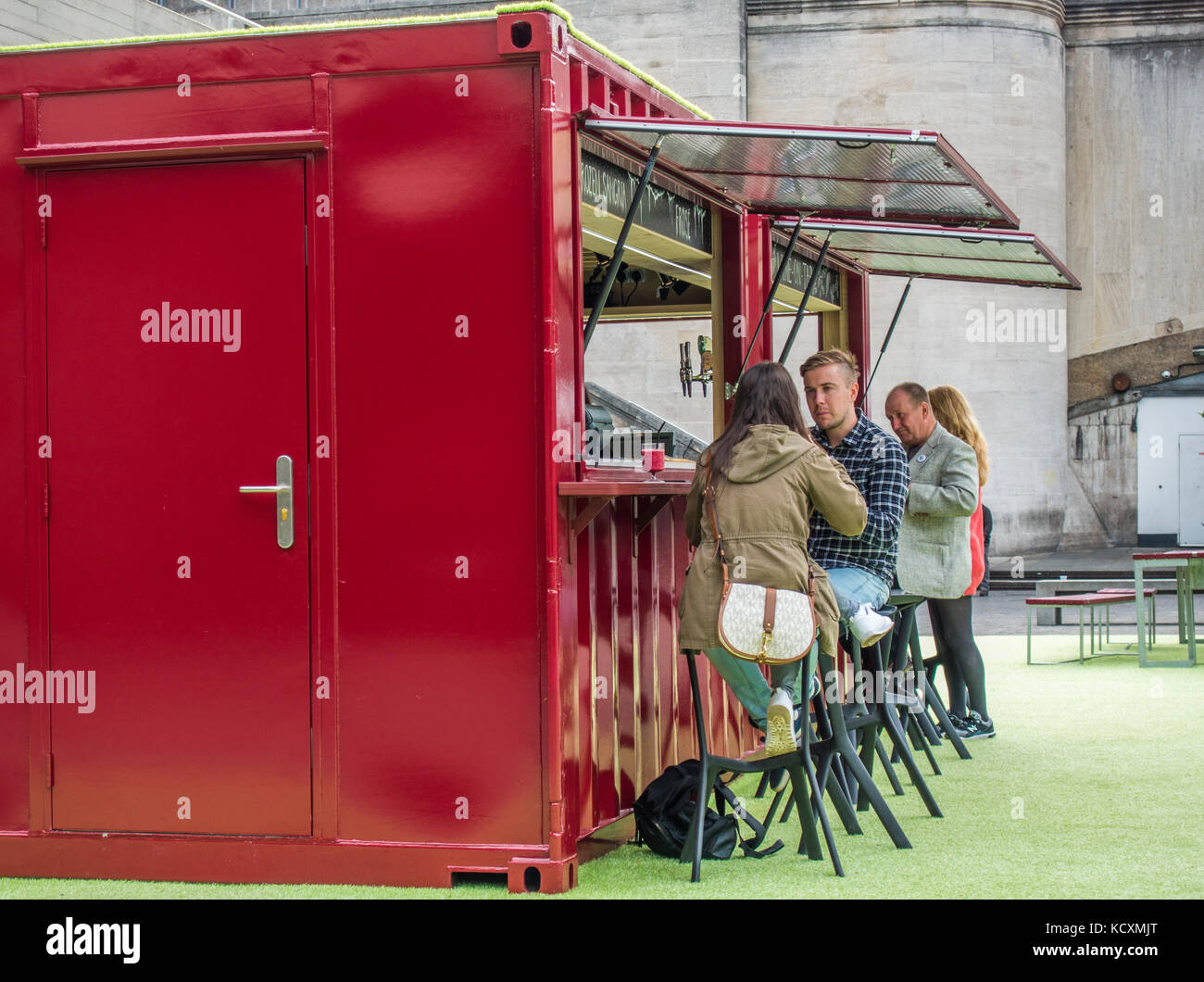 People drinking at a wine bar, made from a converted shipping container and painted red. Outside on the South Bank, London, England, UK. Stock Photo