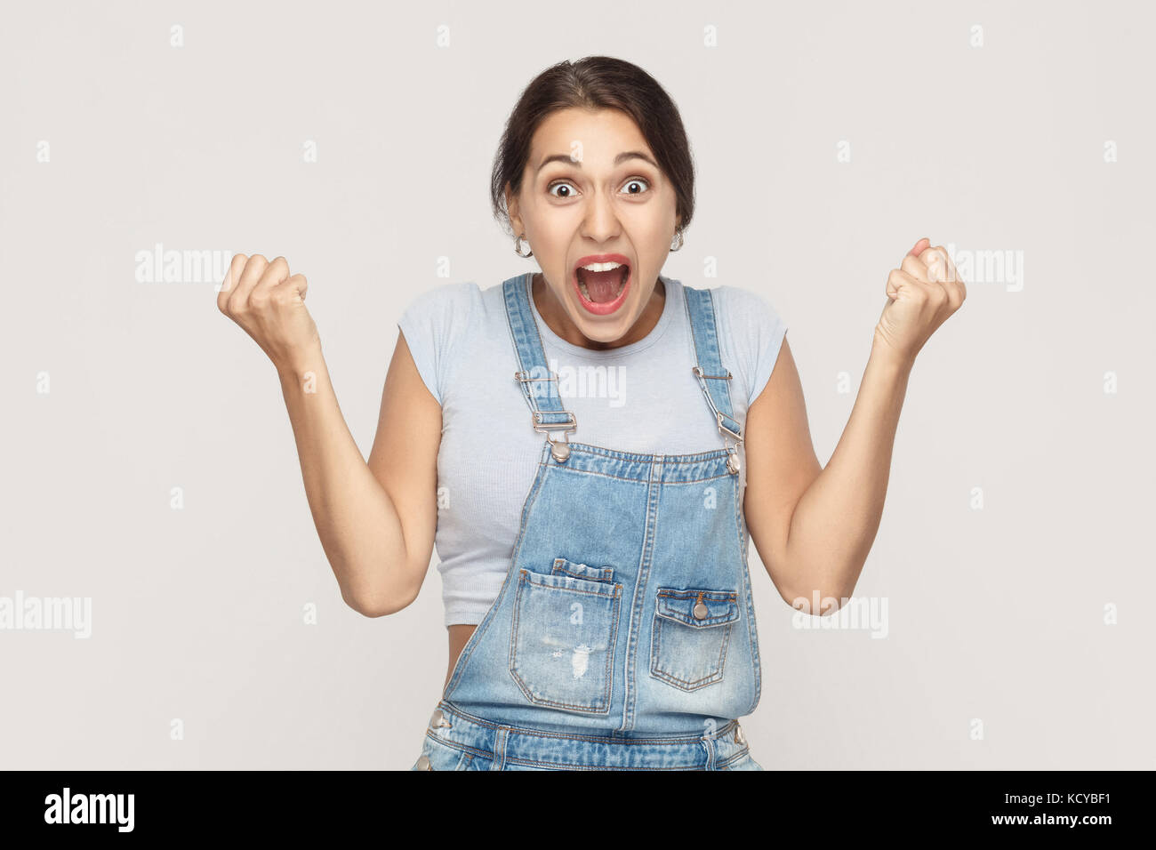 Happiness woman in denim overalls rejoicing for his success. Isolated on gray background. Indoor, studio shot Stock Photo