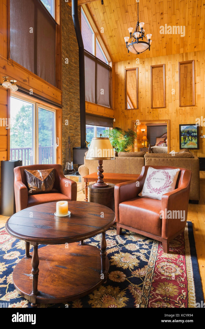 Round wooden coffee table and reddish-brown leather sitting chairs in reading area of the great room with cathedral ceiling inside a milled cottage style flat log profile home, Quebec, Canada Stock Photo
