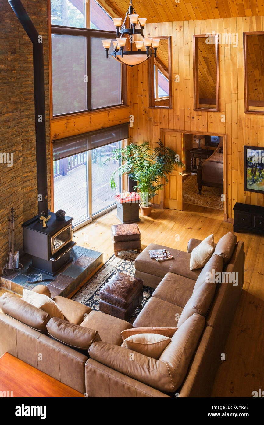 Toip view of tan leather sectional sofa and wood burning stove in living room area of the great room with cathedral ceiling inside a milled cottage style flat log profile home, Quebec, Canada Stock Photo