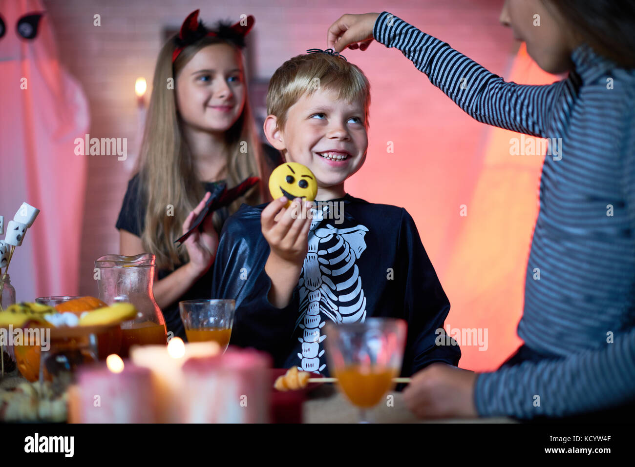 Portrait of children wearing Halloween costumes playing in decorated room during party, girl putting toy spider on top of cute little boys head Stock Photo