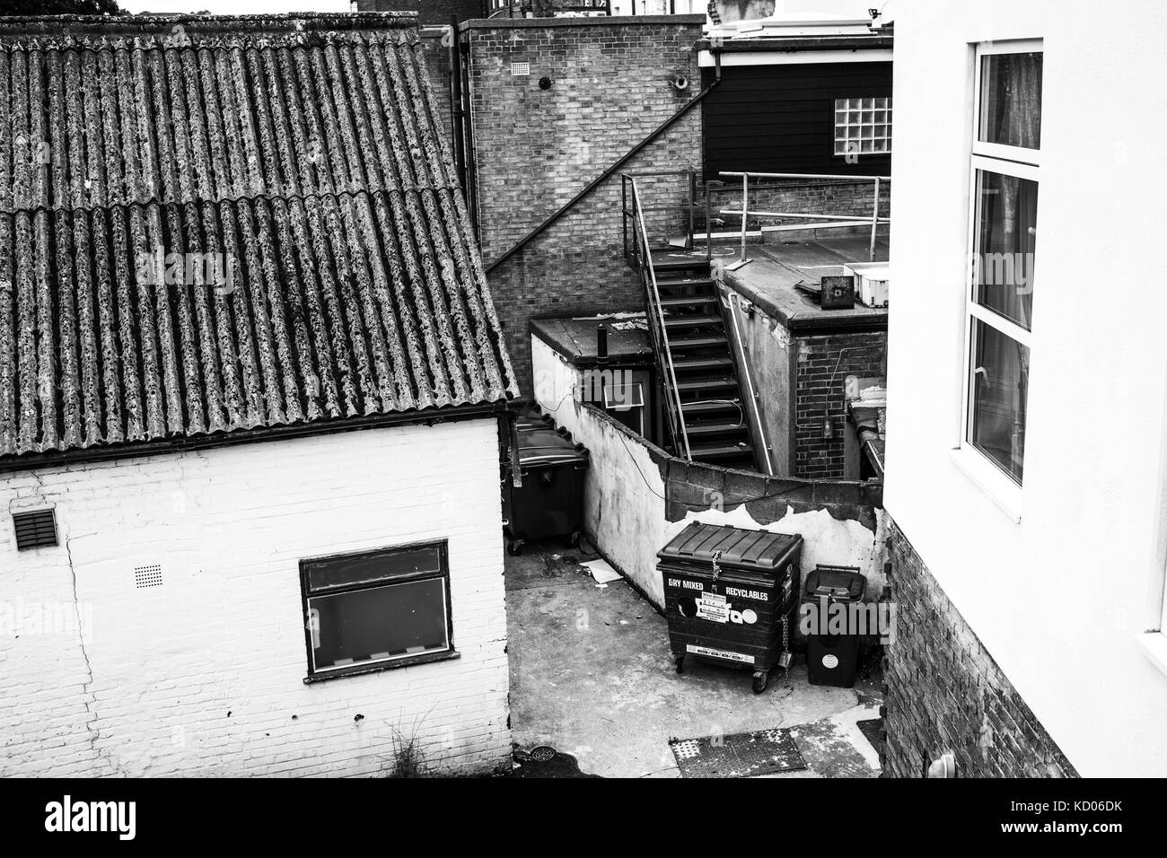Black and White Monochrome Image of an Urban Alleyway With Wheely Bins in Disrepair and Brick Buildings with No People Stock Photo