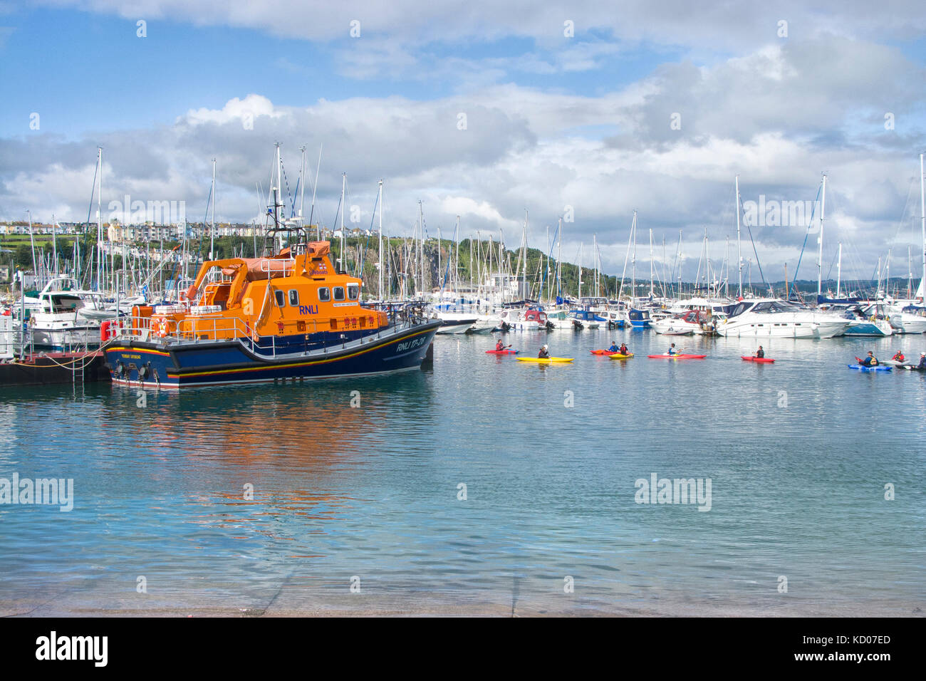 Summer in Brixham Harbour with children kayaking alongside the Torbay Lifeboat. Yachts and motor boats, pontoon and Brixham town in background. Stock Photo