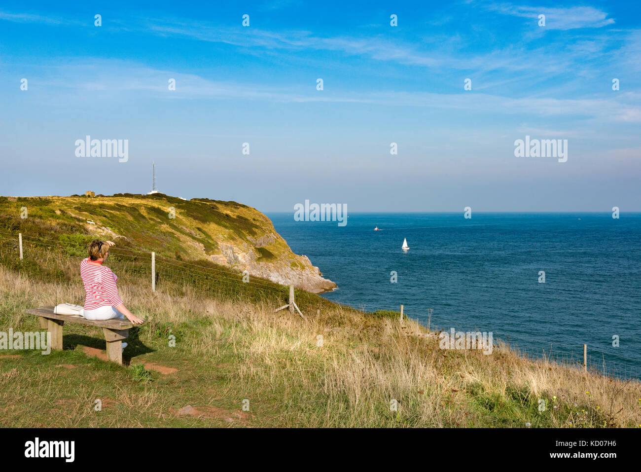 Lady takes a rest from walking the South West Coast Path at Berry Head, Brixham, Devon looking out to sea at a single yacht on sunny summer's day. Stock Photo