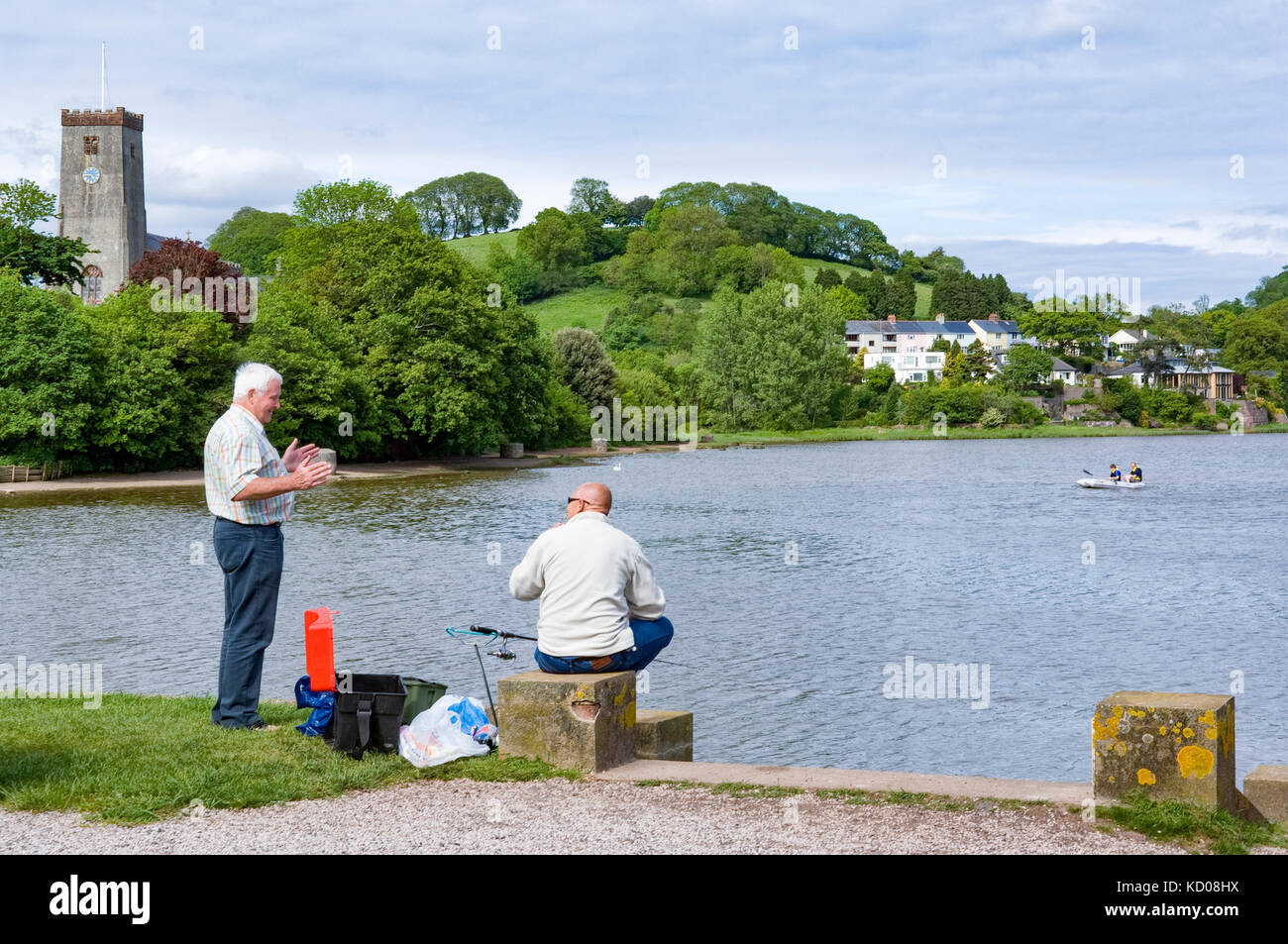 Fishing on the River Dart at Stoke Gabriel, Devon seated man and standing man talking. boat on the river with church tower and houses across the water Stock Photo
