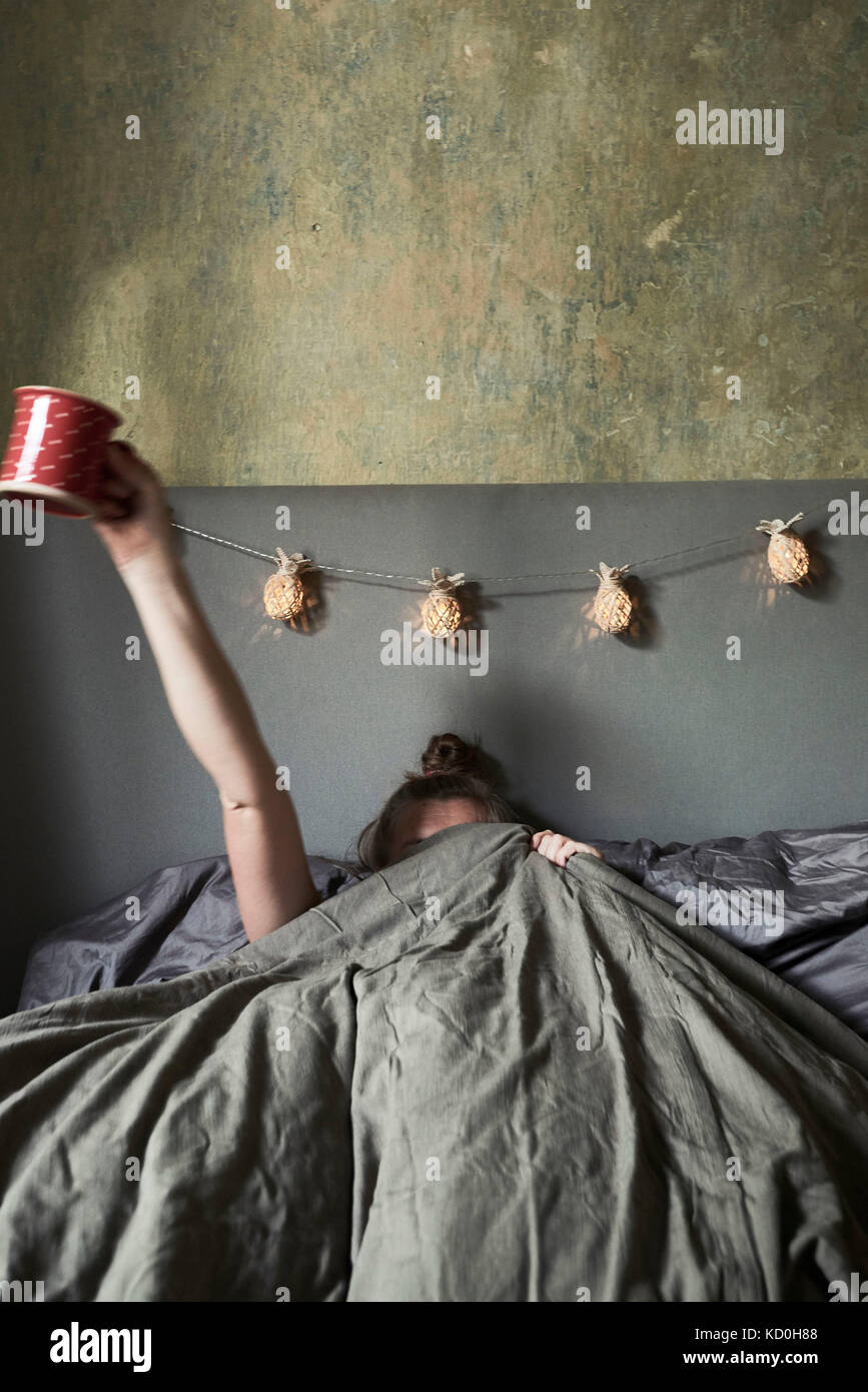 Woman in bed, hiding under covers, holding mug in air Stock Photo