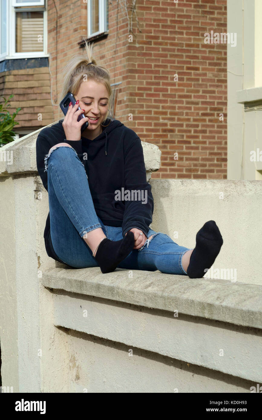 young female student sitting on a wall outside of her accommodation engrossed in conversation on her smartphone england uk Stock Photo