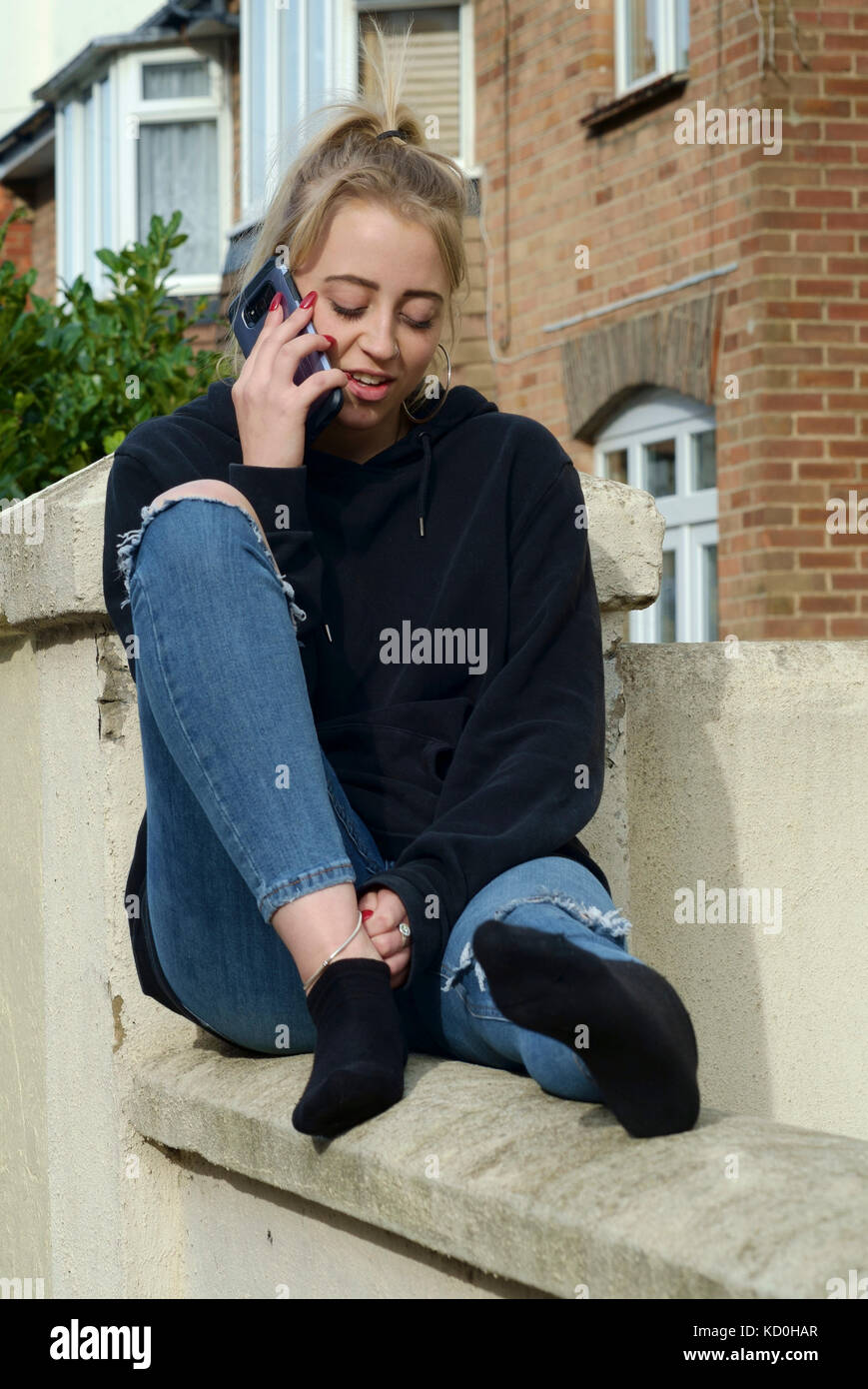 young female student sitting on a wall outside of her accommodation engrossed in conversation on her smartphone england uk Stock Photo