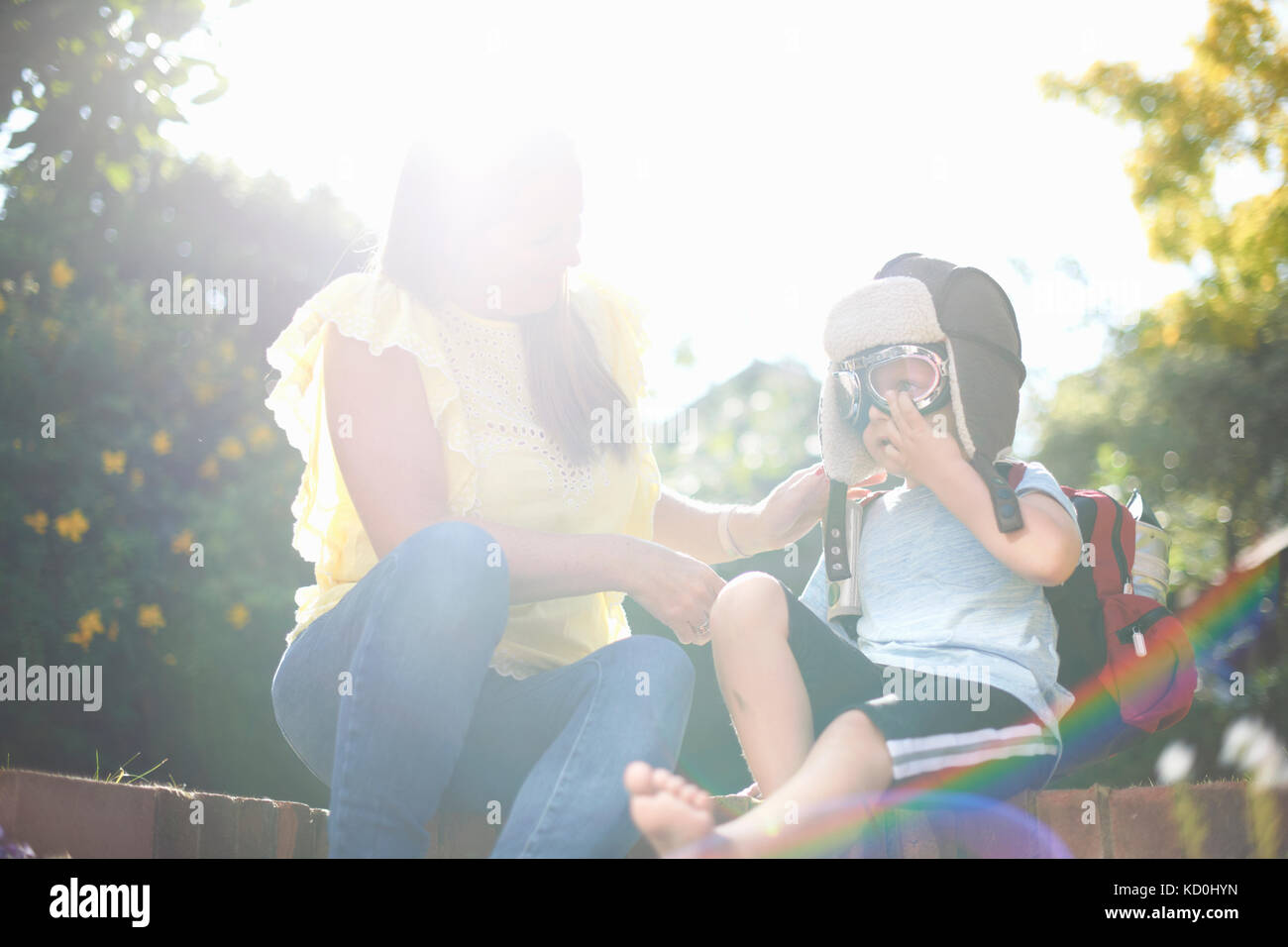 Boy with mother in sunlit garden putting on pilot flying goggles Stock Photo