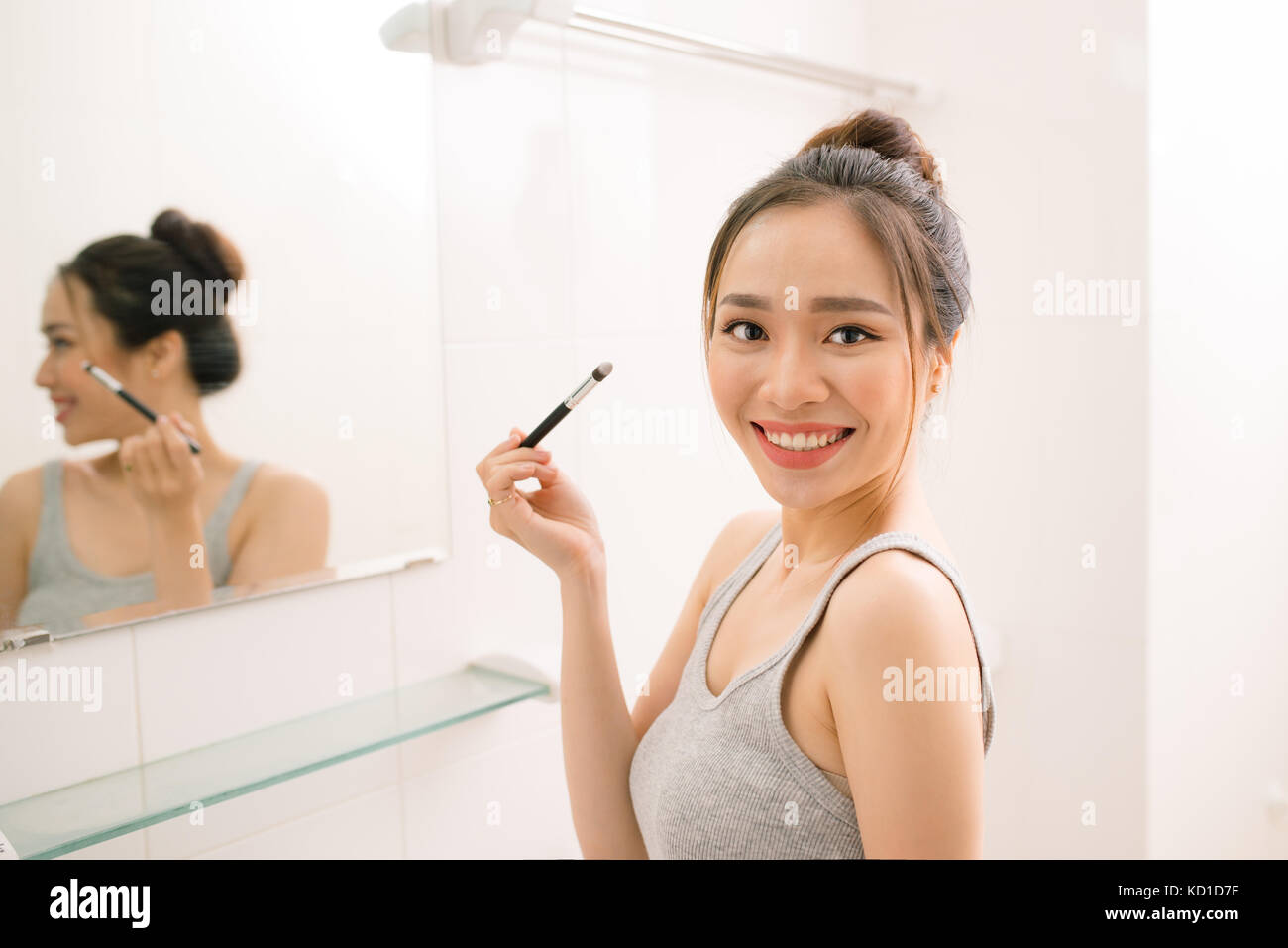 Smiling young woman looking to mirror doing makeup at home bathroom Stock Photo