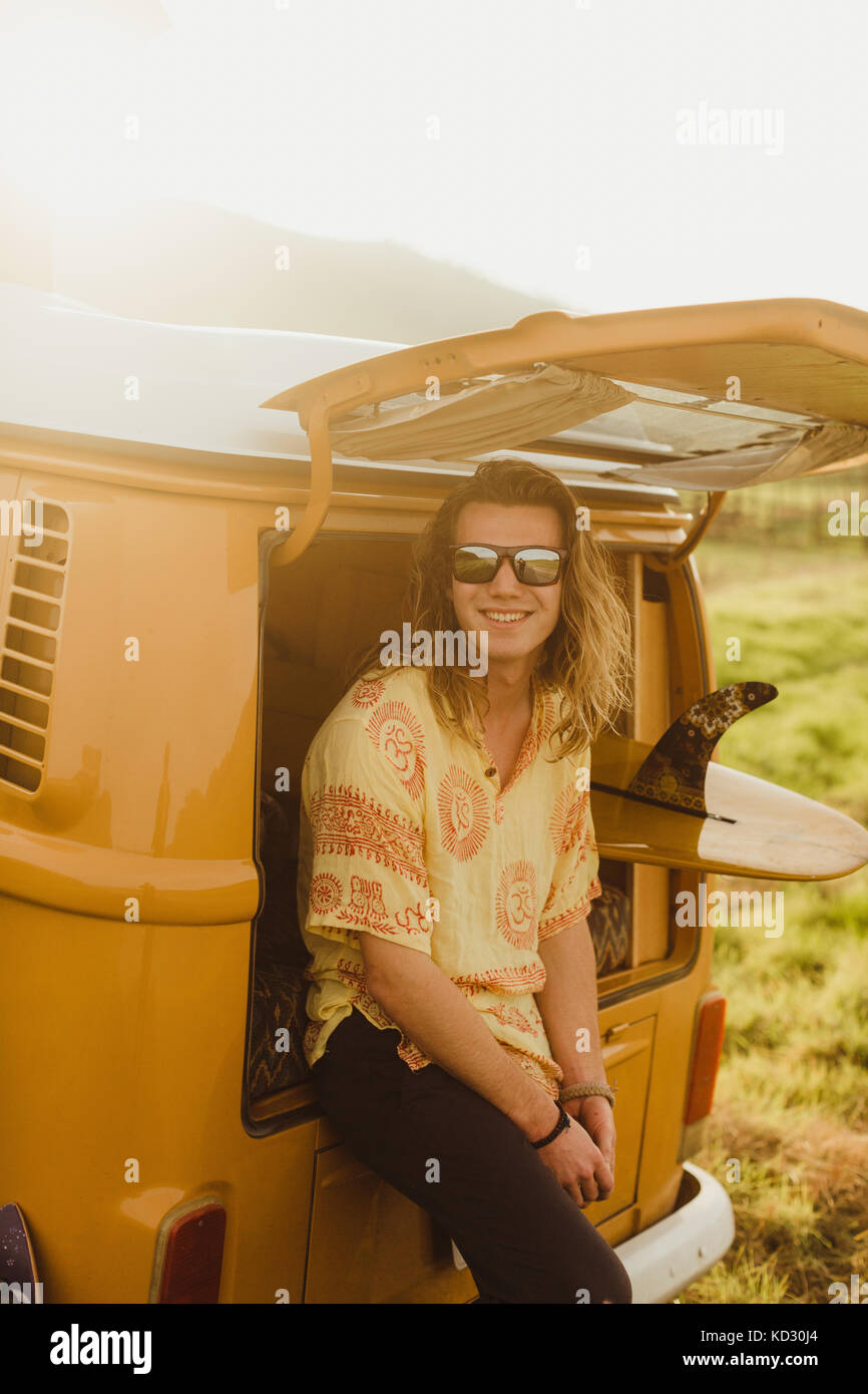 Portrait of young male surfer with long hair leaning against vintage recreational vehicle, Exeter, California, USA Stock Photo