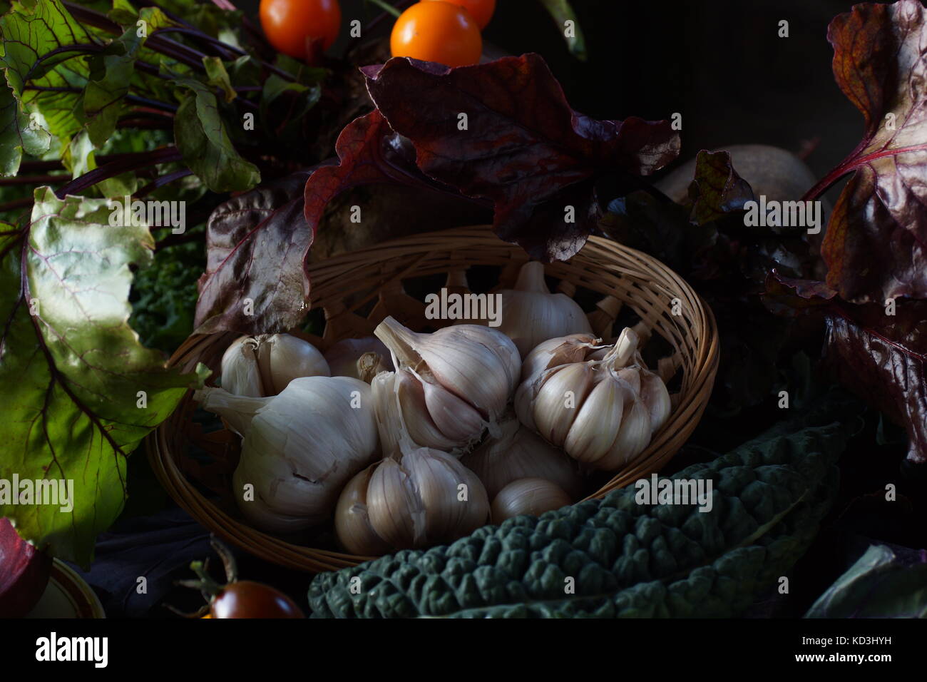 Still life: basket with garlic, tomatoes, kale leaves  and Brussels red cabbage  among the greens on the table. Stock Photo