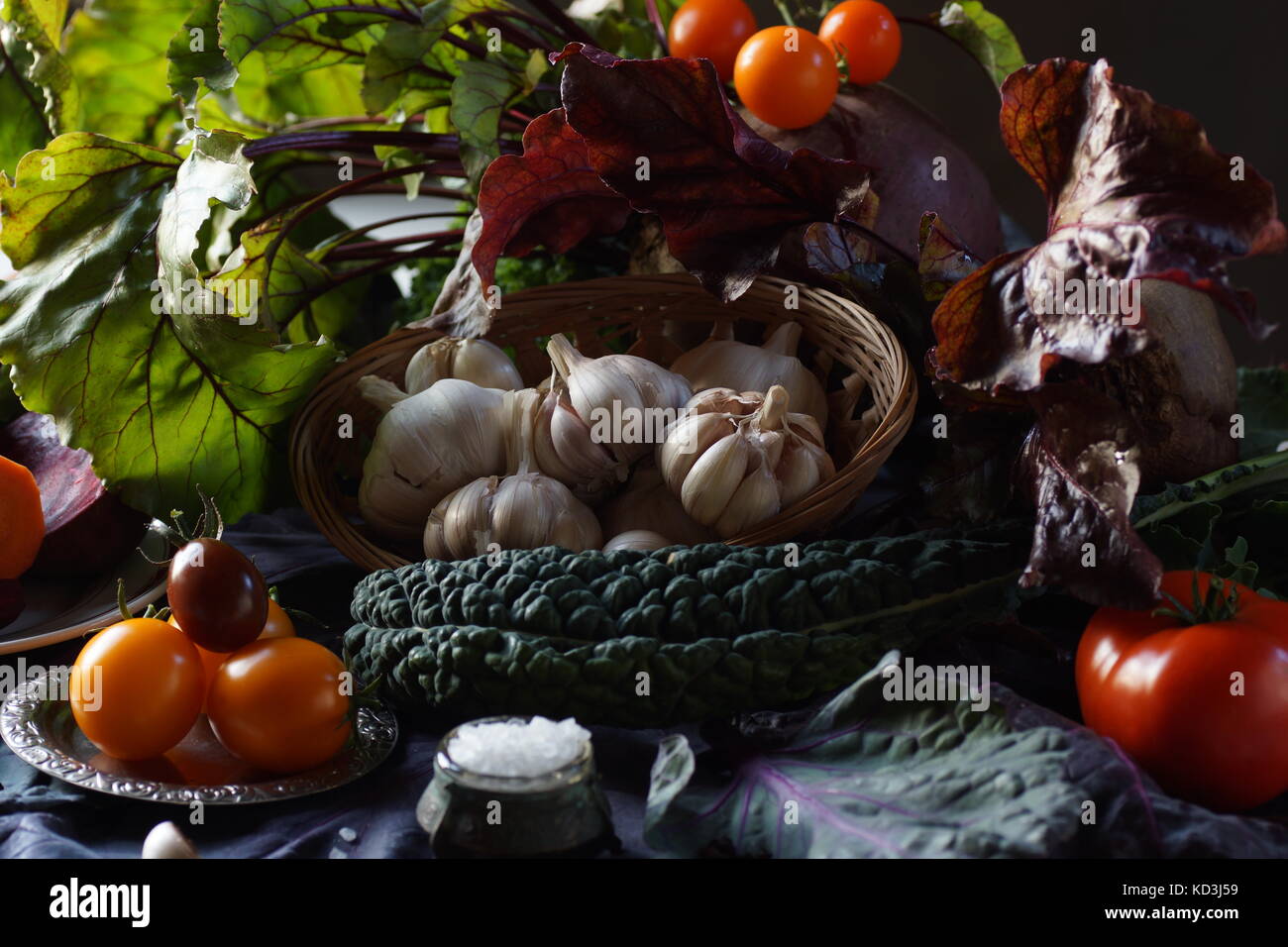 Still life: basket with garlic, tomatoes, kale leaves, Brussels red cabbage and salt among the greens on the table. Stock Photo