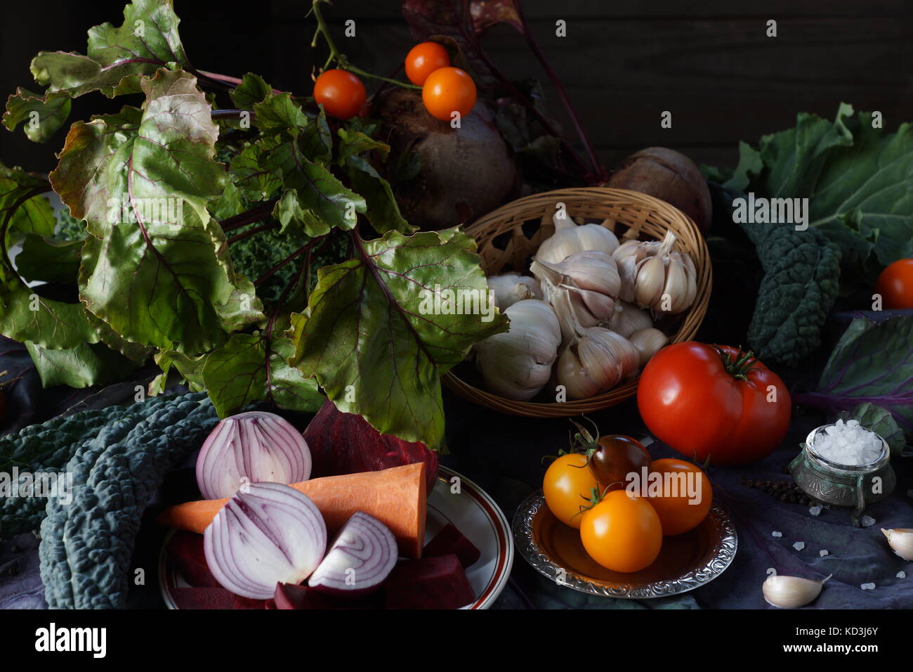 Still life: basket with garlic, tomatoes, kale leaves, red onions, carrots, Brussels red cabbage and salt among the greens on the table. Stock Photo
