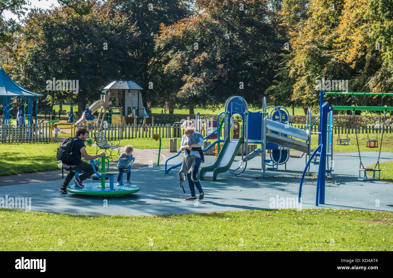 Young parents and their two small children in a playground / play area, on a warm sunny day, early autumn. Gunnersbury Park, West London, England, UK. Stock Photo