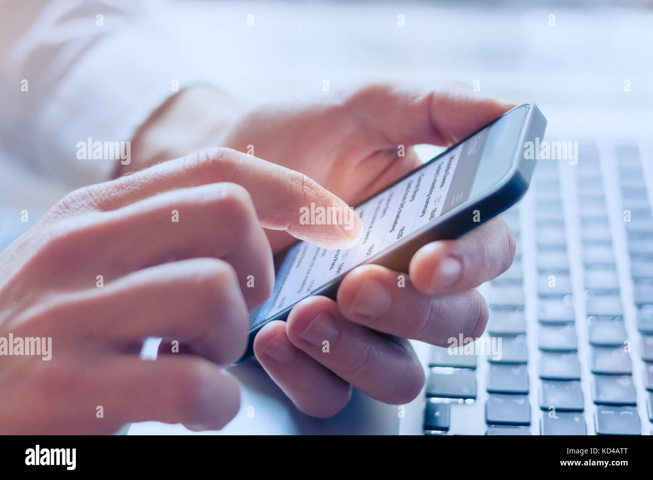 Manager sending email to contact his business team with smartphone, close-up on hand, laptop in background Stock Photo