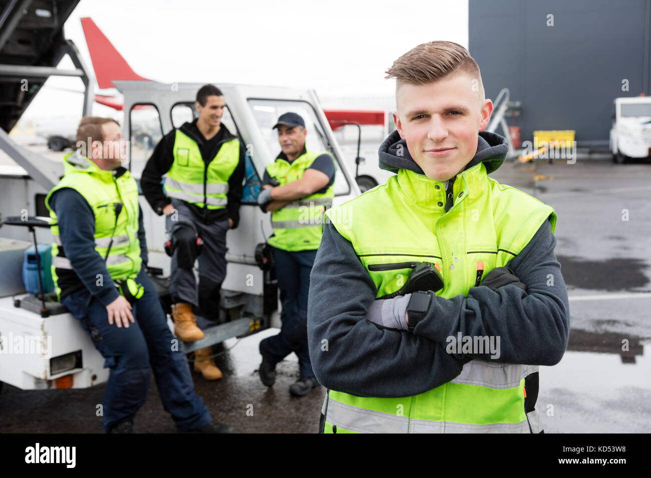 Portrait of confident male worker with arms crossed standing while colleagues communicating at gangway on runway Stock Photo