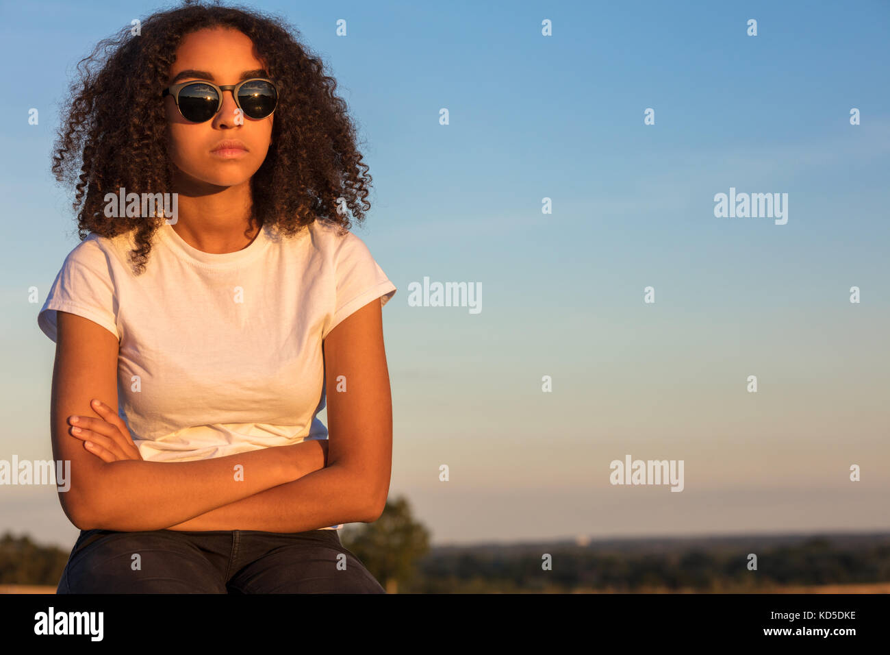 Beautiful mixed race African American girl teenager female young woman outside wearing sunglasses looking sad depressed or thoughtful at sunrise or su Stock Photo