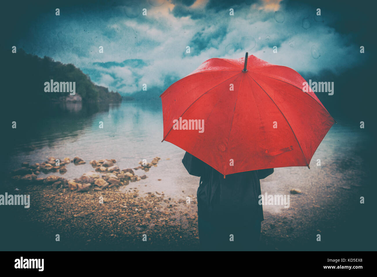 Woman with red umbrella contemplates on rain in front of a lake. Sad and lonely female person looking into distance. Grunge editing with dirt, noise,  Stock Photo
