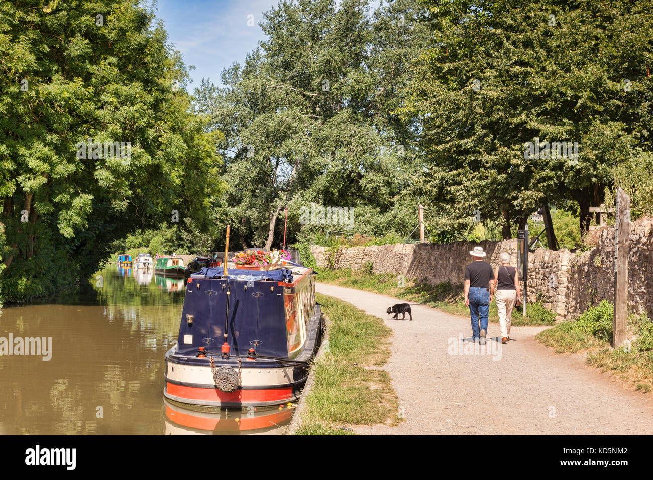 7 July 2017: Bradford on Avon, Somerset, England, UK - Couple with dog walking on the tow path beside a row of narrowboats moored along the banks of t Stock Photo