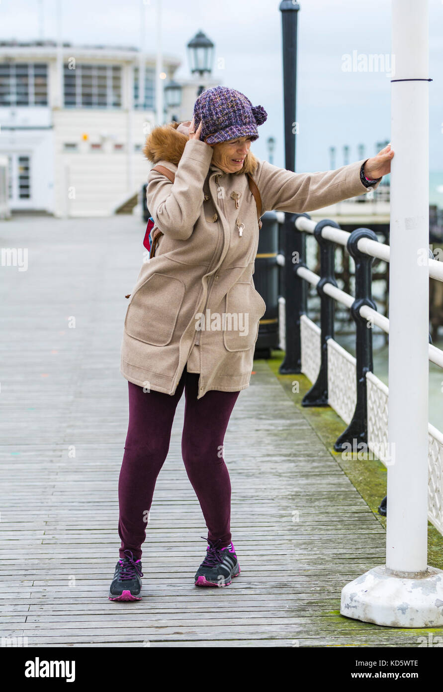 Woman holding onto a post while struggling to walk in the wind on Worthing Pier in Worthing, West Sussex, England, UK. Stock Photo