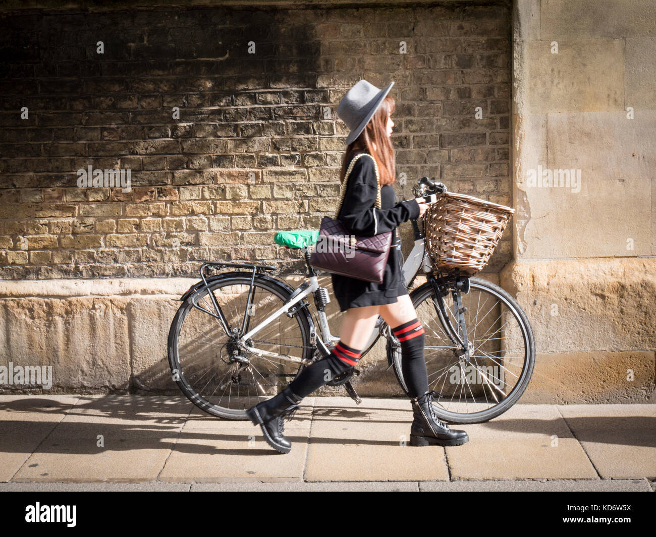 A Japanese woman wearing long socks walks past a bicycle with a basket leaning up against the wall of Sidney Sussex College part of Cambridge Universi Stock Photo