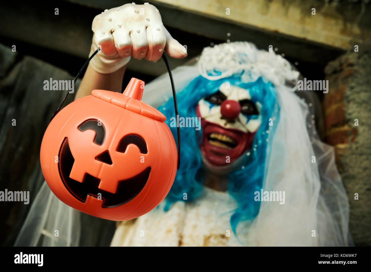 closeup of a scary evil clown wearing a bride dress, with a veil and a diadem, at the door of a scary house holding a treat basket in the shape of a j Stock Photo