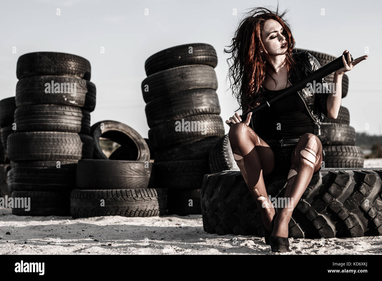 Disheveled redhead woman sitting with a baseball bat at the background of an old tires Stock Photo