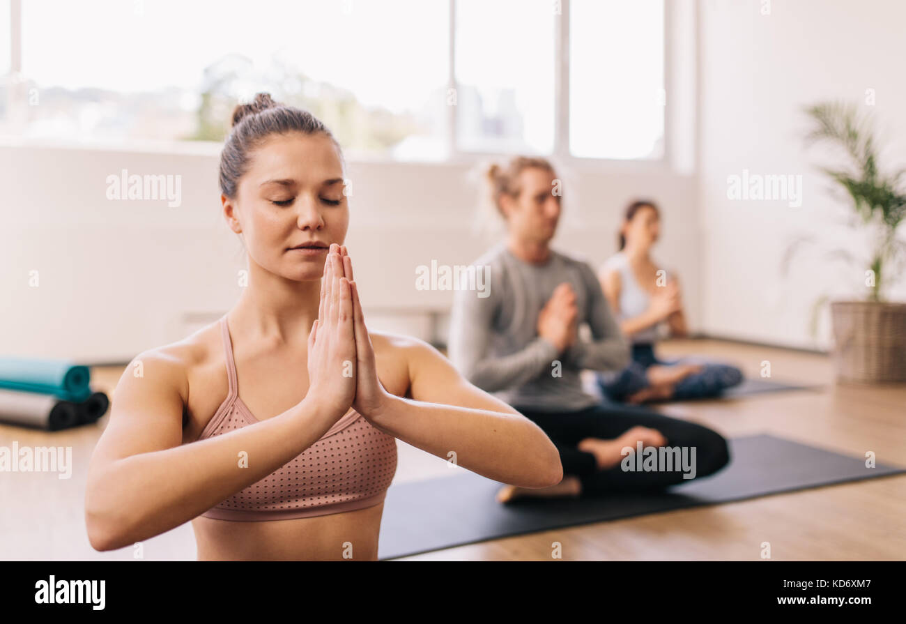 Young woman practicing yoga in gym class in fitness center with people in background. People meditating at health club. Stock Photo