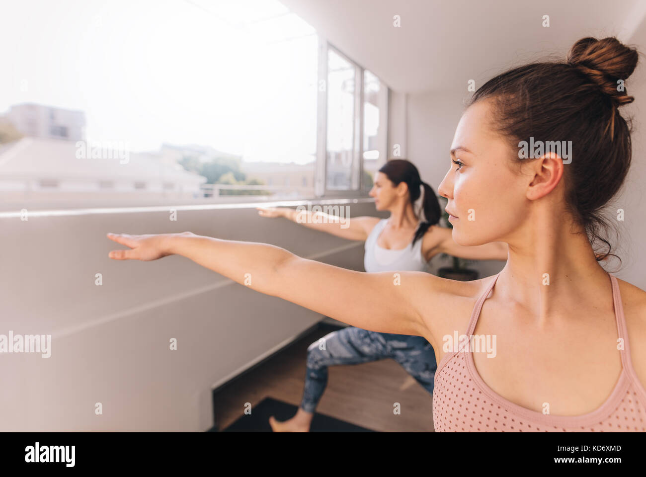 Women doing yoga in warrior pose at studio. Young fitness females working out indoors. Stretching yoga workout in gym class. Stock Photo